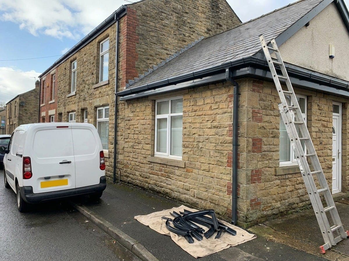 Newly fitted dark grey guttering and downpipe on a Lancashire stone house, with a plain van and ladder set up for a replacement