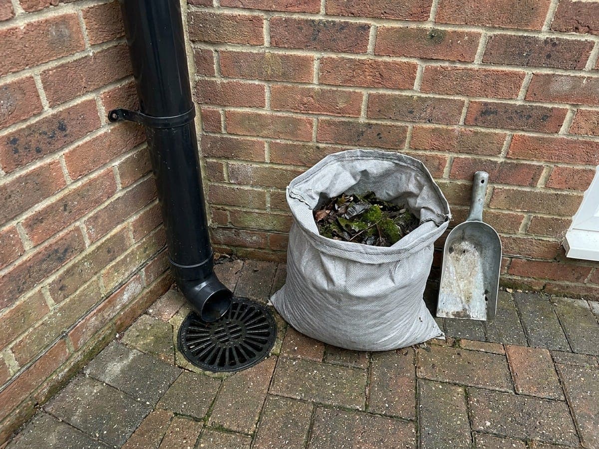A cleared downpipe outlet and gully grate beside a sack of bagged gutter debris and a scoop