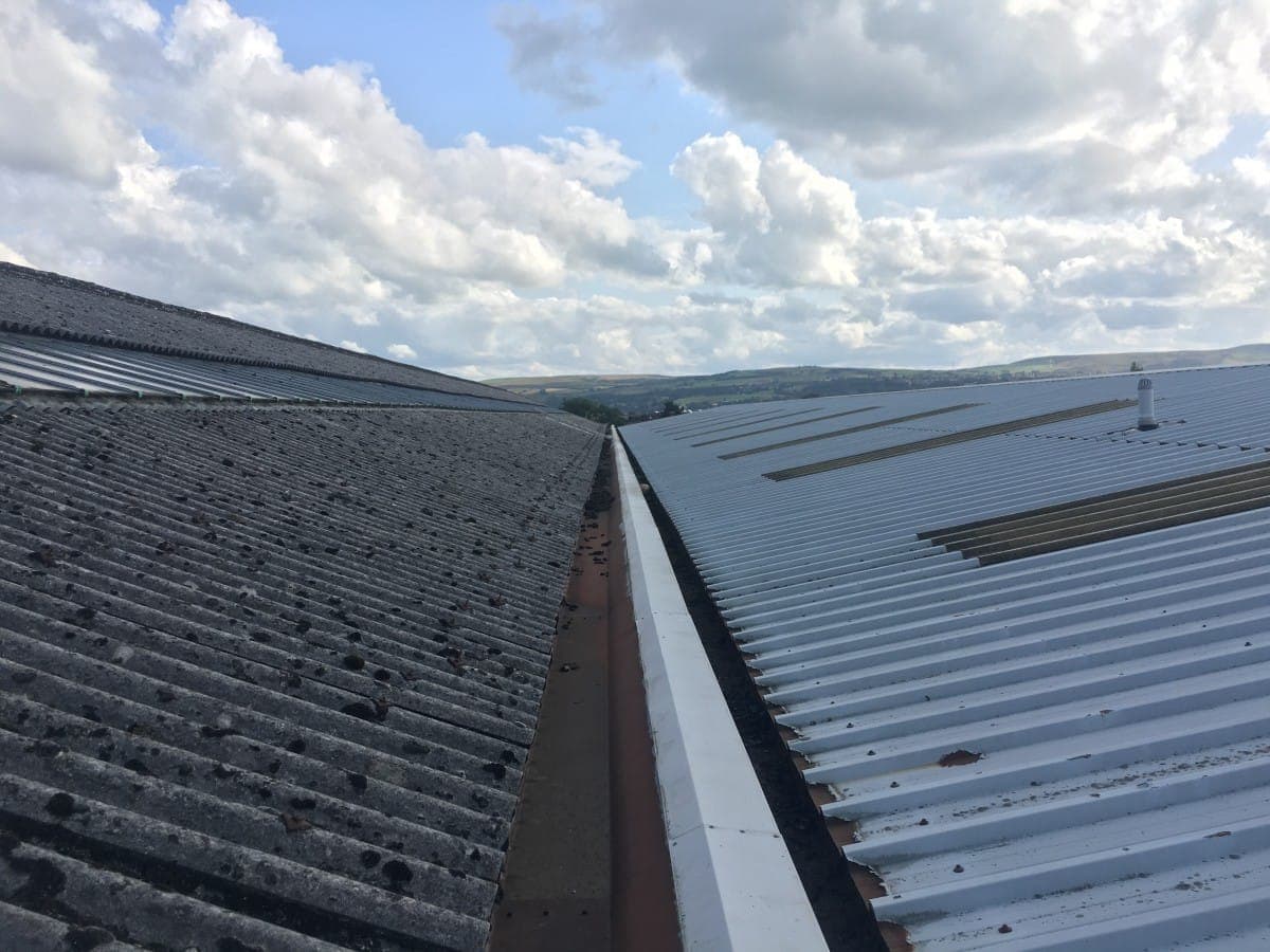A metal box gutter running between two large corrugated commercial roofs, with the Lancashire hills behind